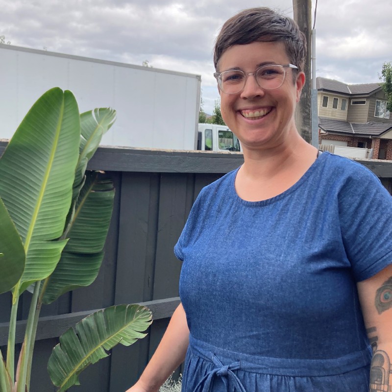 Anita Conlon stands beside a broad flat leaf plant in front of a dark grey steel fence. She is wearing glasses and a blue t-shirt and smiling broadly.